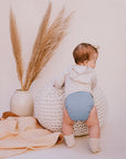 Baby in a blue all in one cloth diaper standing next to a textured chair and vase with dried plants on a light background