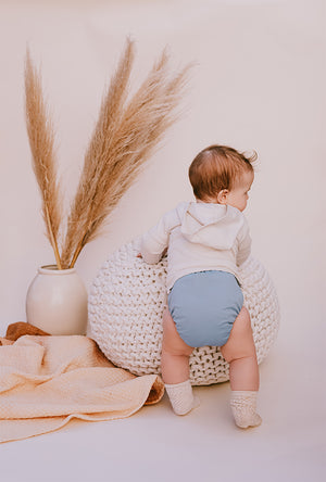 All-In-One Cloth Diaper, baby sitting on a crochet pouf next to decorative pampas grass.